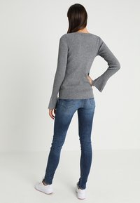 Kvinna som står med ryggen vänd, iklädd en grå ribbad långärmad tröja, blå tajta jeans och vita sneakers mot en neutral bakgrund.