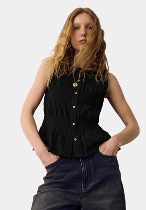 Kvinde med langt krøllet blondt hår iført en ærmeløs sort bluse med knapper og mørke højtaljede jeans, hænderne i lommerne foran en ensfarvet baggrund.