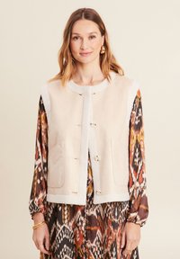 Beige Wildlederweste mit einem flauschigen weißen Kragen und Knopfverschluss, ausgestattet mit zwei Fronttaschen und einem geraden Saum. Trägt man über einem gemusterten Kleid.