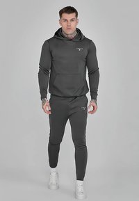 Ciemnoszara sportowa bluza z kapturem z przednią kieszenią i logo, w komplecie z dopasowanymi zwężanymi spodniami dresowymi. Gładka tkanina, dopasowany krój i białe trampki.