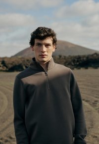 Πουλόβερ fleece σε σκούρο γκρι, διαθέτει ψηλό γιακά και φερμουάρ μέχρι το ένα τέταρτο. Μαλακή υφή με εφαρμοστή σχεδίαση, φορεμένο από ένα άτομο σε εξωτερικό χώρο.