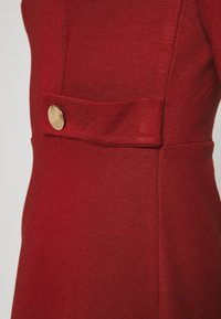 Czerwona sukienka o dopasowanym fasonie, z teksturowanej tkaniny, z akcentem złotego guzika po boku. Czyste linie i prosta sylwetka.