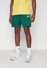 Zelene mrežaste atletske hlače z rumenim logotipom Adidas, v kombinaciji z rumeno mrežasto majico in sivimi atletskimi čevlji, oblečeni z belimi nogavicami.