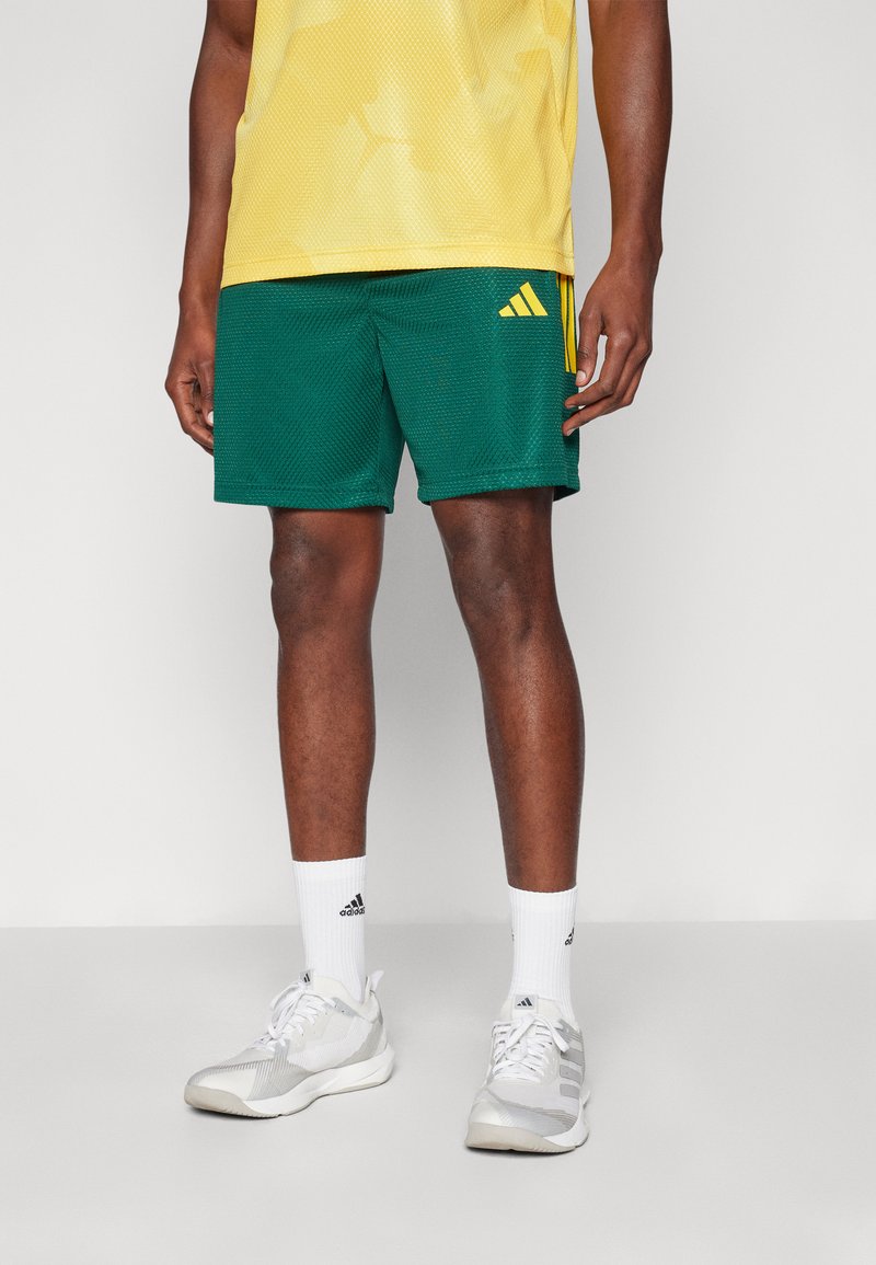 Zelene mrežaste atletske hlače z rumenim logotipom Adidas, v kombinaciji z rumeno mrežasto majico in sivimi atletskimi čevlji, oblečeni z belimi nogavicami.