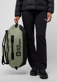Green and black duffel bag with logo, made of durable material. Person in black pants and jacket stands nearby, holding the bag.