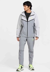 Chaqueta Nike de color gris claro con capucha, detalles en negro y bolsillos con cremallera, combinada con pantalones gris claro a juego y zapatillas negras con lima.