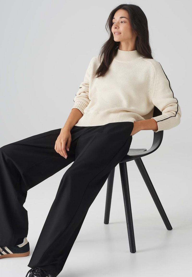 Cremefarbener Strickpullover mit hohem Kragen, kombiniert mit schwarzen weit geschnittenen Hosen. Schwarze und weiße Sneakers runden das Outfit ab.