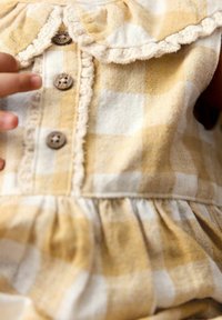 Gelbes und weißes kariertes Stoffkleid mit rüschigem Kragen, Holzknöpfen und gerafftem Taillenbereich. Weiche Textur, Baumwollmaterial.