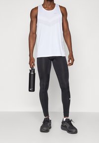 Mies, jolla on päällään valkoinen hihaton urheilu paita ja mustat leggingsit, mustat urheilukengät jalassa, ja hän pitää mustaa urheiluvesipulloa oikeassa kädessään.