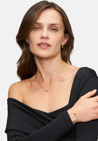 Goldenes herzförmiges Pendant-Halsband und passende Ohrringe, getragen von einem Model in einem schwarzen schulterfreien Oberteil, mit glatten Texturen und zarten Ketten.