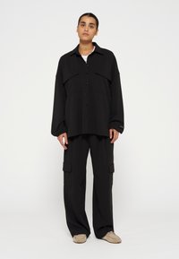 Schwarze, übergroße Button-down-Bluse mit zwei Brusttaschen, kombiniert mit passenden weit geschnittenen Hosen. Weicher Stoff mit lässiger Passform.