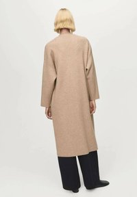 Persona con capelli biondi corti che indossa un lungo cappotto beige, pantaloni neri a gamba larga e scarpe nere, in piedi con la schiena rivolta su uno sfondo neutro.