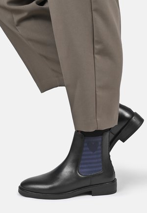 Black leather ankle boots with navy blue elastic side panels worn with beige pants on a light background.