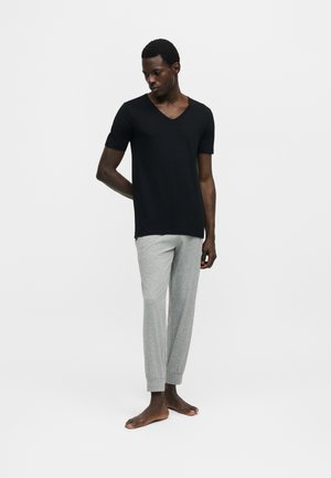 Bărbat stând desculț, purtând un tricou negru cu mâneci scurte și gât în V și pantaloni de trening gri deschis pe un fundal alb simplu.