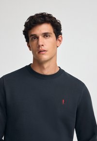 Sudadera gris oscura con cuello redondo y un pequeño logo de golfista rojo en el lado izquierdo del pecho. Tejido suave con un corte relajado.