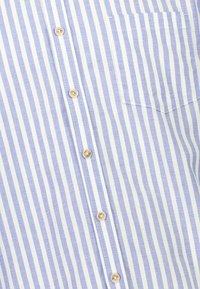 Vaaleansininen ja valkoinen raidallinen nappipaita. Puuvillamateriaali, jossa on kultaiset napit ja yksi rintatasku. Pystyraidat koko paidan matkalla.