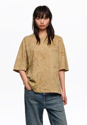 Mujer joven con cabello largo y oscuro, vistiendo una camiseta beige extragrande con estampado floral y vaqueros azules holgados, de pie con una mano en el bolsillo.