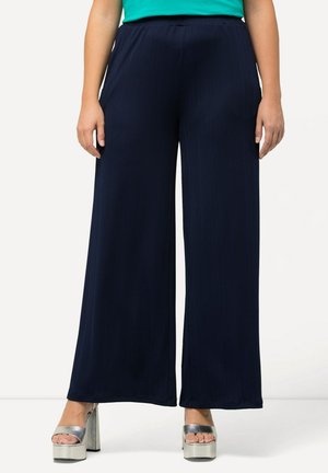 Kobieta ubrana w granatowe spodnie o szerokich nogawkach, turkusowy top i srebrne sandały na platformie, stojąca na tle jasnego, jednolitego tła.