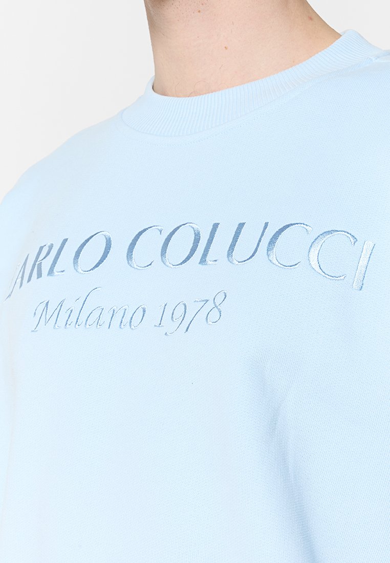 Lichtblauwe sweatshirt met ribbelboordkraag. Bevat reliëftekst "CARLO COLUCCI Milano 1978" in een contrasterende glanzende afwerking.