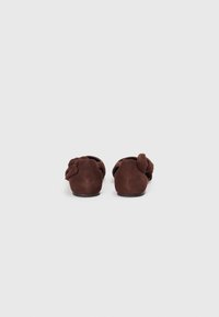 Brown suede slip-on shoes with a rounded toe, featuring a bow accent at the back and a smooth texture. Flat heel design.