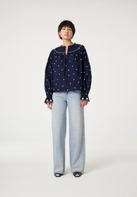 Marinebluse mit weißer Blumenstickerei, Puffärmeln und einem Schlüsselloch-Ausschnitt, kombiniert mit hellblauen weiten Jeans und schwarzen Sandalen.