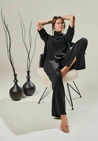 Schwarzer Strickpullover mit hohem Kragen, kombiniert mit lockeren schwarzen Hosen. Das Modell sitzt auf einem schwarzen Stuhl neben zwei dunklen Vasen und dünnen Ästen.