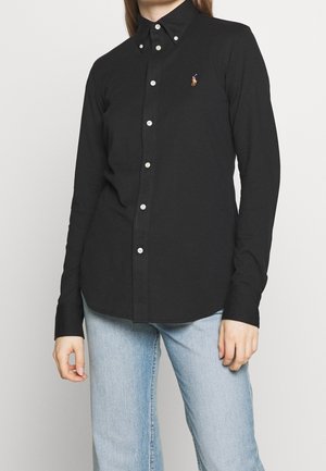 Persoon draagt een zwart, knoopsluitend overhemd met lange mouwen en een klein geborduurd logo op de borst, gecombineerd met lichtblauwe spijkerbroek.