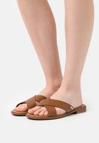 Sandales en cuir marron avec des brides croisées, semelles plates et finition texturée. Semelle intérieure légèrement rembourrée, affichant la forme naturelle du pied.