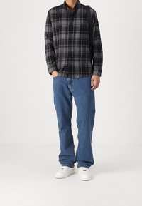 Muž v černém a šedém kostkovaném zapínacím triku, volných modrých džínech, bílých teniskách a s černou taškou přes rameno na jednobarevném pozadí.