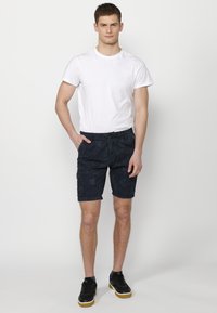 Fiatal férfi áll, kezében a zsebében, fehér pólót, sötét mintás sortot és fekete tornacipőt viselve, egy sima fehér háttér előtt.