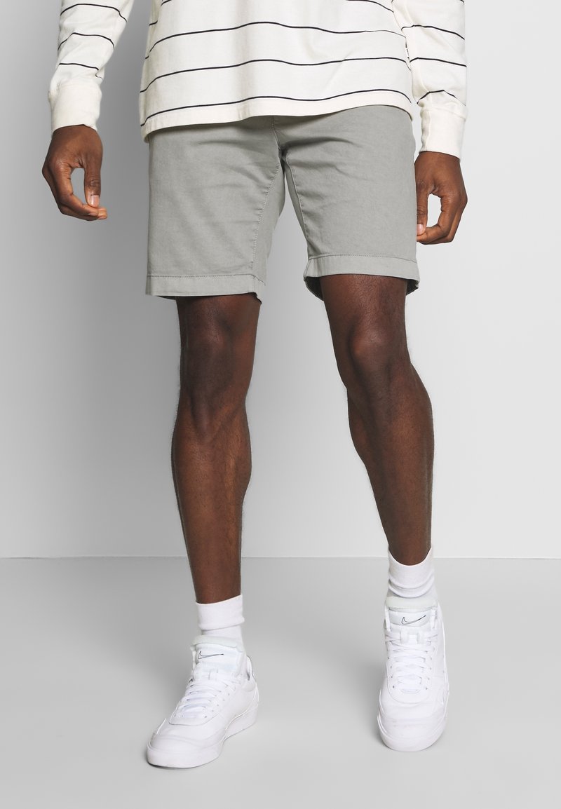 Uomo in pantaloni corti grigi, camicia a maniche lunghe bianca a righe, calze bianche e sneaker bianchi Nike, in piedi su uno sfondo neutro.