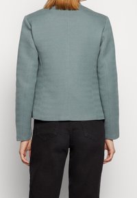 Přiléhavá bunda v tlumeném modro-zeleném herringbone vzoru, texturovaná látka, dlouhé rukávy a rovný střih v pase.