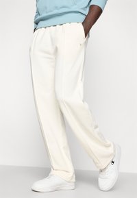 Ανοιχτό μπεζ φόρμα με ελαστική μέση, πλαϊνές τσέπες και διακριτικές λεπτομέρειες ραφής, σε συνδυασμό με λευκά αθλητικά παπούτσια.