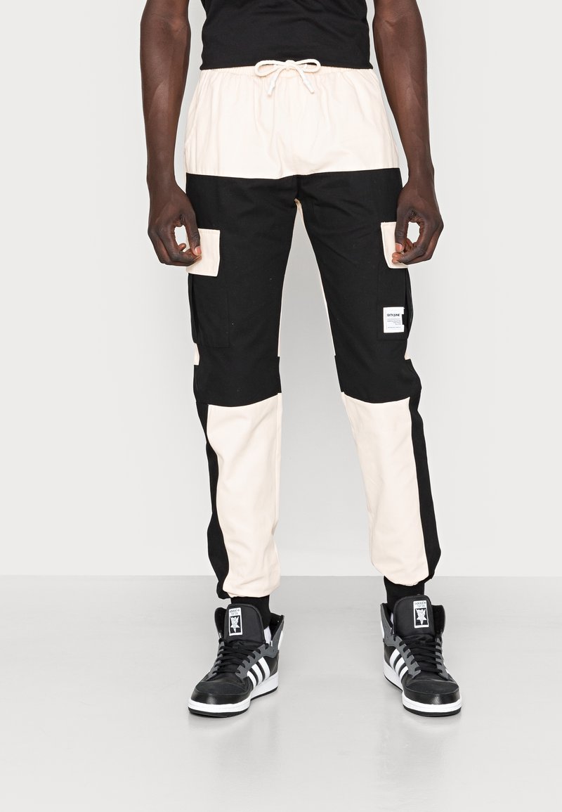 Homme portant un pantalon cargo bicolore noir et blanc avec taille à cordon et des baskets montantes noires avec des rayures blanches.