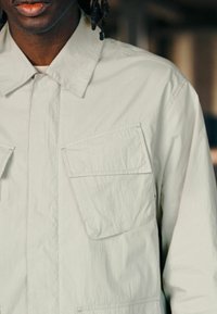 Camisa ligera de color gris claro, con diseño de botones, dos bolsillos frontales y un cuello clásico. Textura suave y ajuste casual.