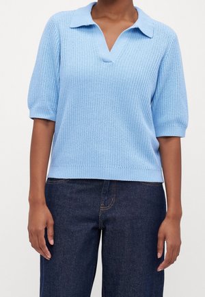 Personne portant un polo en maille bleu clair à manches courtes, associé à un jean en denim bleu foncé, debout devant un fond uni.