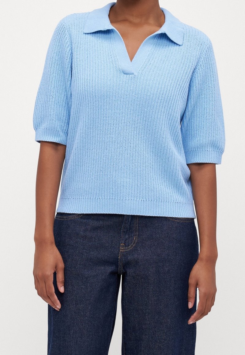 Personne portant un polo en maille bleu clair à manches courtes, associé à un jean en denim bleu foncé, debout devant un fond uni.