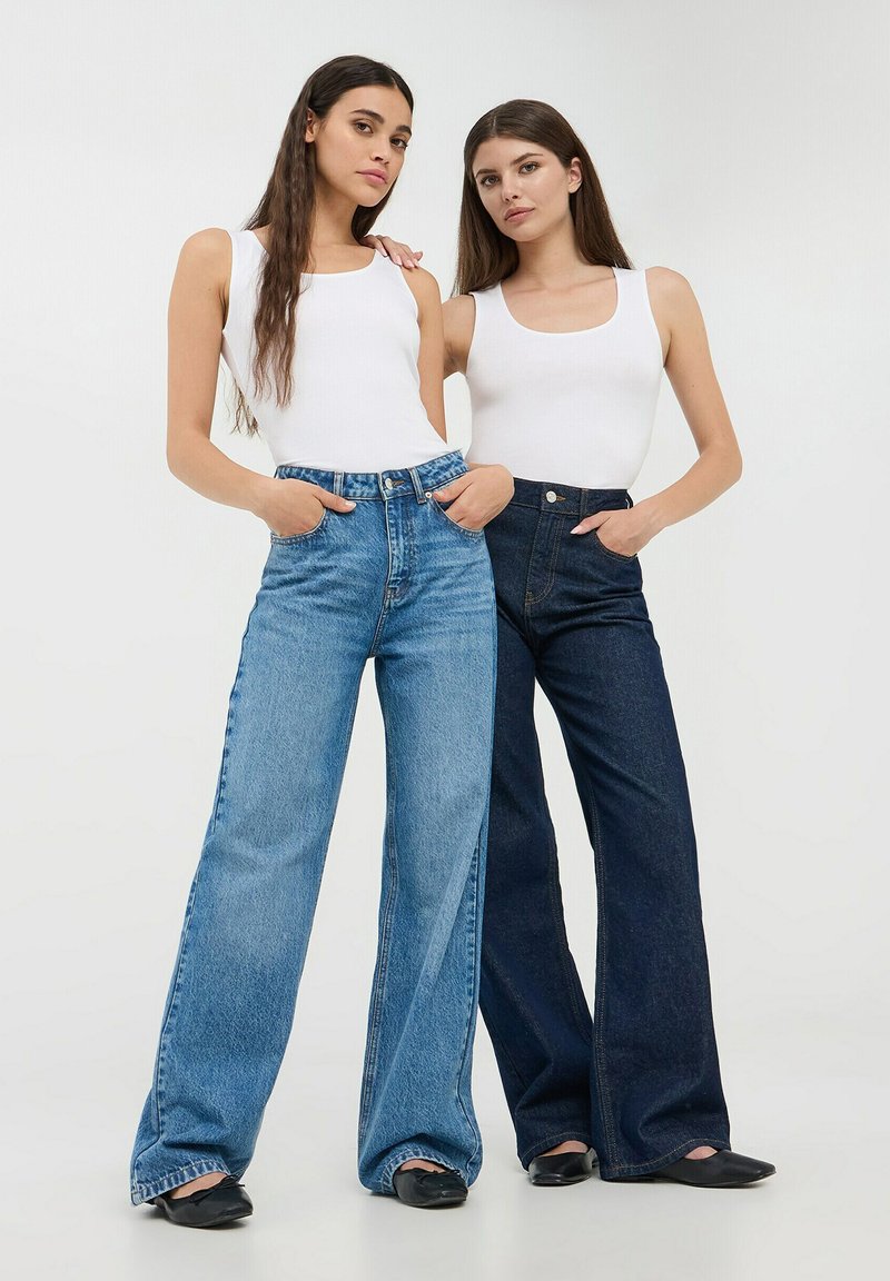 Jeans a gamba larga di un blu chiaro con tasche frontali a sinistra; jeans a gamba larga di un blu scuro a destra, entrambi abbinati a canotte bianche.