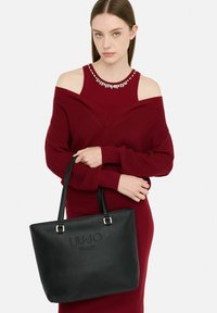 Czarna skórzana torba typu tote z wytłoczonym logo, złotym metalowym wykończeniem i dwoma paskami na ramię. Gładka tekstura, prostokątny kształt i pojemny design.