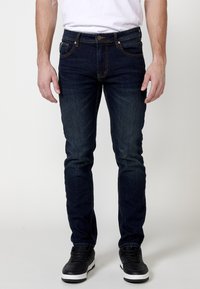 Jeans in denim blu scuro con vestibilità slim, cinque tasche, cuciture a contrasto e chiusura con bottone, abbinati a sneakers alte nere.