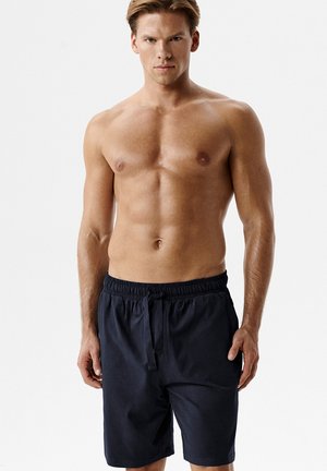 Wysportowany mężczyzna o jasnej karnacji i krótkich blond włosach, ubrany w czarne szorty z troczkiem, stojący na jednolitym jasnym tle.