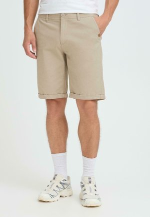 Beige Baumwollshorts mit umgeschlagenen Säumen, Seitentaschen und Knopfverschluss vorne. Kombiniert mit weißen Sportsocken und hellen Sneakers.