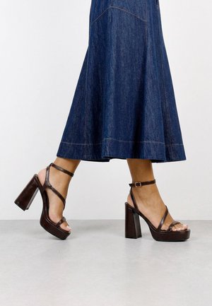 Mujer caminando con sandalias de plataforma de tacón alto de color marrón oscuro con correas en el tobillo, vistiendo una falda larga de mezclilla.