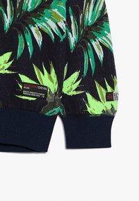 Pull avec une base noire présentant de grands motifs de feuilles verts et blancs. Poignets et ourlet côtelés en bleu marine. L'étiquette indique "Retour Denim, Deluxe."