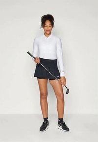 Fehér, hosszú ujjú golf póló és fekete szoknya, golfütőt tart. A póló galléros; a szoknya pliszírozott. Fekete sportcipő.