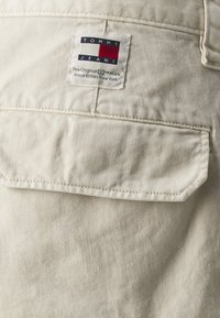 La tela de mezclilla de algodón beige presenta una etiqueta de parche visible con rectángulos rojos, blancos y azules, cosidos con líneas rectas a través del bolsillo.