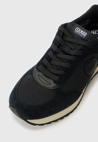 Schwarze Sneaker aus einer Mischung von Wildleder und Mesh-Materialien, runde Zehenpartie, dicke Sohle und Logodetail auf der Zunge. Die Schnürsenkel sind flach und schwarz.