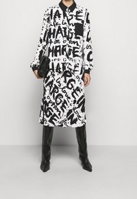 Outfit in bianco e nero con un audace motivo di testo; include una camicia con colletto e una gonna coordinata, abbinata a stivali neri lucidi fino al ginocchio.