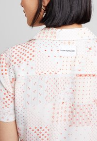 Short-sleeve blouse in lightweight fabric, featuring a white background with red geometric and floral patterns. Label at the back: "CALVIN KLEIN JEANS."