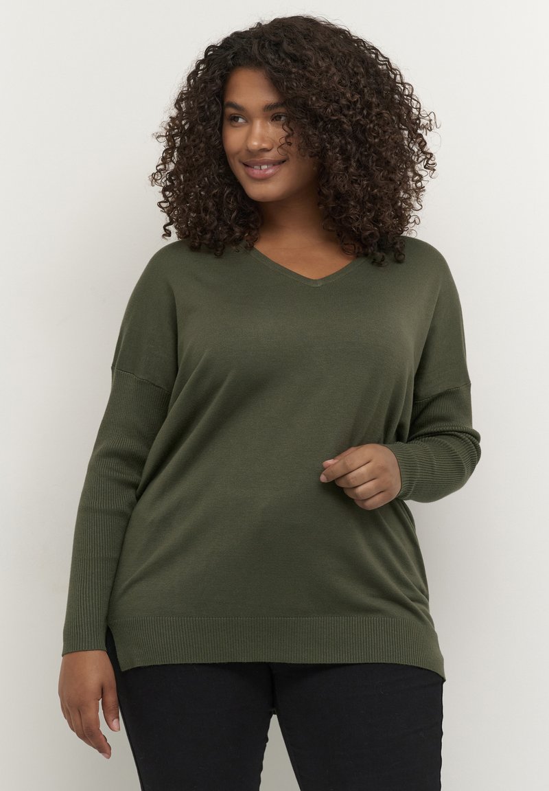 Maglione verde oliva a scollo a V con texture a coste e spacchi laterali. Maniche lunghe, vestibilità comoda. Indossato con jeans scuri. Tessuto morbido e casual.